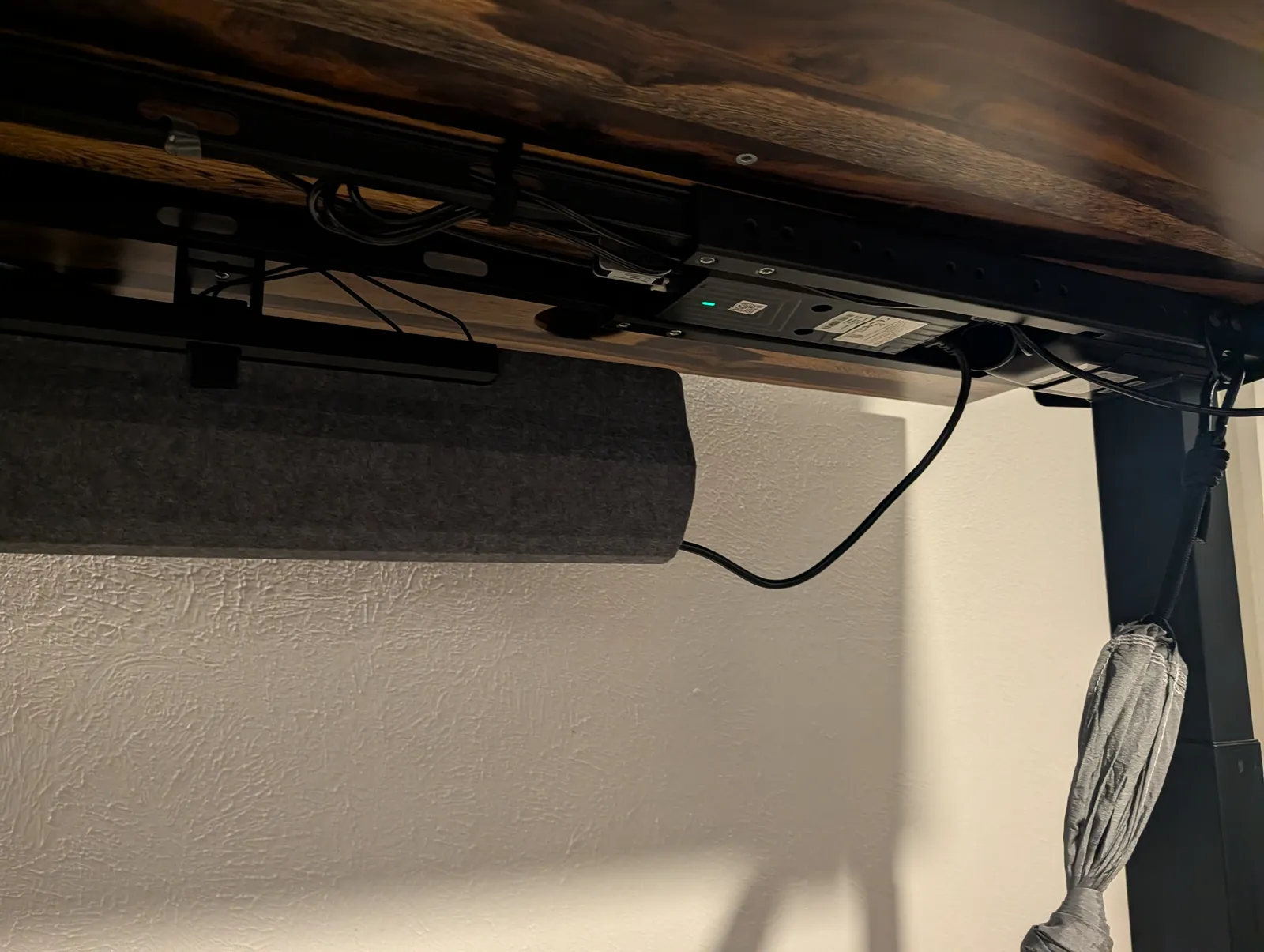 Under the back of the desk: the gray felt FlexMount cable pouch slung the length of the frame, with the mountable surge protector screwed to the underside beside it, indicator light on.